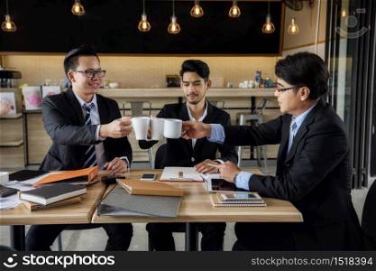 Asian entrepreneur businessman team toast or clink glasses of hot coffee after getting agreement of project in coffee cafe. Work at home Happy business teamwork working in cafeteria.