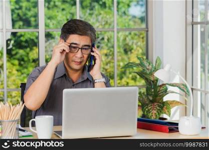 Asian executive senior businessman sitting on desk office he using his mobile phone and talking with somebody, the confident middle aged handsome man using laptop computer at workplace home office