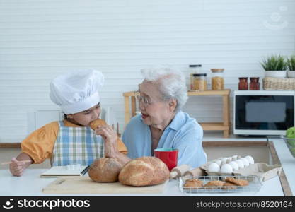 Asian family grandmother and grandchild have fun cooking and baking cookies at home kitchen together, little cute granddaughter with chef hat and apron is eating homemade cookie from Grandma