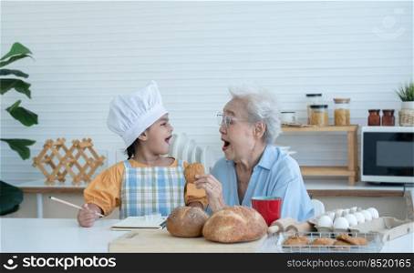 Asian family grandmother and grandchild have fun cooking and baking cookies at home kitchen together, little cute granddaughter with chef hat and apron is eating homemade cookie from Grandma