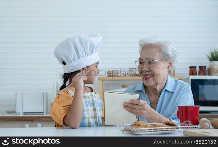 Asian family grandmother and grandchild have fun cooking at home kitchen together, little cute granddaughter with chef hat and apron is writing down recipe and how to make cookie from Grandma