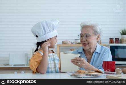 Asian family grandmother and grandchild have fun cooking at home kitchen together, little cute granddaughter with chef hat and apron is writing down recipe and how to make cookie from Grandma