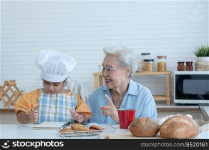 Asian family grandmother and grandchild have fun cooking at home kitchen together, little cute granddaughter with chef hat and apron is writing down recipe and how to make bread or cookie from Grandma