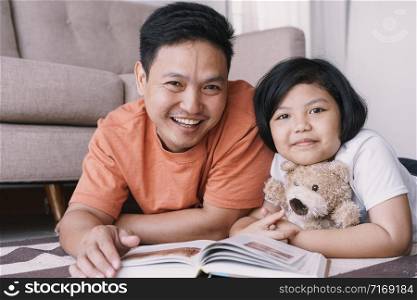 Asian father and daughter are reading book while lying on the carpet in the living room at her home. The cute little girl hugs the doll and smile happily with his father.