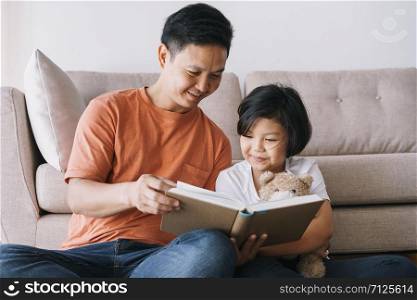 Asian father and daughter reading book while sitting in the living room at her home.