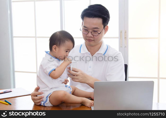 Asian father feeding milk by glass for adorable daughter at home office multitask working and parenthood, father taking care cute girl and running small business entrepreneur logistic with laptop