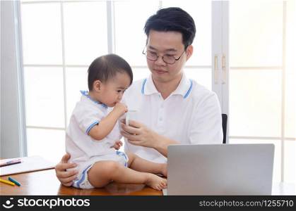 Asian father feeding milk by glass for adorable daughter at home office multitask working and parenthood, father taking care cute girl and running small business entrepreneur logistic with laptop
