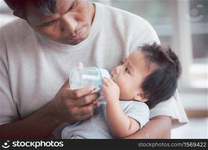 Asian father holding and feeding her baby boy with milk from baby bottle.