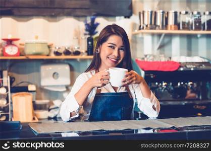 Asian female barista making cup of coffee. Young woman holding white coffee cup while standing behind cafe counter bar in restaurant background. People lifestyles and Business occupation concept.