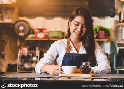 Asian female barista making cup of coffee. Young woman holding white coffee cup while standing behind cafe counter bar in restaurant background. People lifestyles and Business occupation concept.