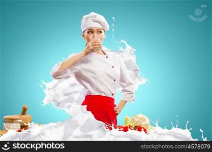 Asian female cook against milk splashes. Asian female cook against milk splashes in red apron against color background drinking milk