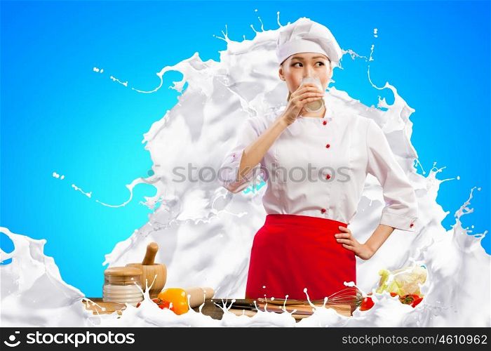 Asian female cook against milk splashes. Asian female cook against milk splashes in red apron against color background drinking milk
