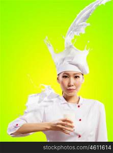 Asian female cook against milk splashes. Asian female cook against milk splashes in red apron against color background holding glass of milk