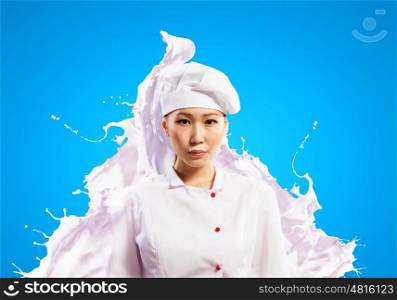 Asian female cook against milk splashes. Asian female cook against milk splashes in red apron against color background holding glass of milk