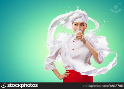 Asian female cook against milk splashes. Asian female cook against milk splashes in red apron against color background drinking milk