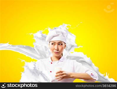 Asian female cook against milk splashes in red apron against color background holding glass of milk