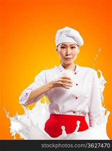 Asian female cook against milk splashes in red apron against color background holding glass of milk