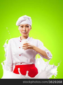 Asian female cook against milk splashes in red apron against color background holding glass of milk