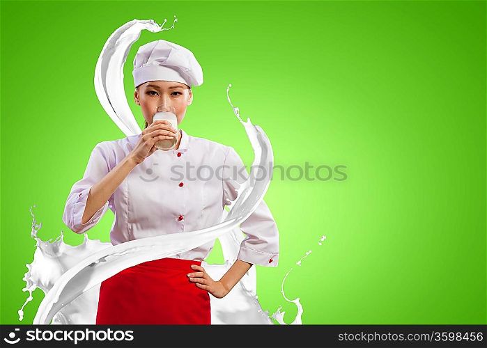 Asian female cook against milk splashes in red apron against color background drinking milk