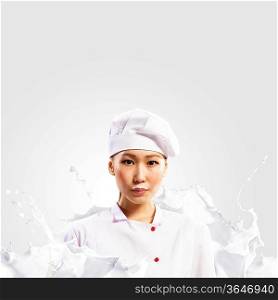 Asian female cook standing against milk splashes in red apron against colo: background