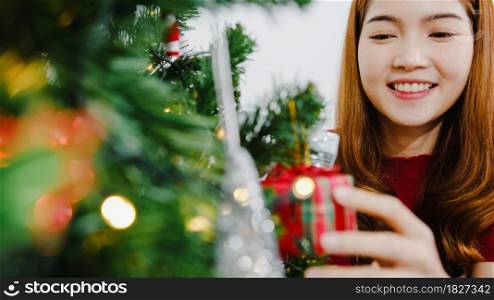 Asian female decorated with ornament on Christmas tree at Christmas and New Year festival in living room at home. Xmas celebration event preparation or winter holidays festival indoor party concept.