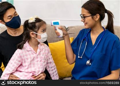 Asian female doctor use infrared thermometer to take temperature to little girl child sit with her dad in living room while doctor visit at home. Home health care delivery and doctor visiting concept.