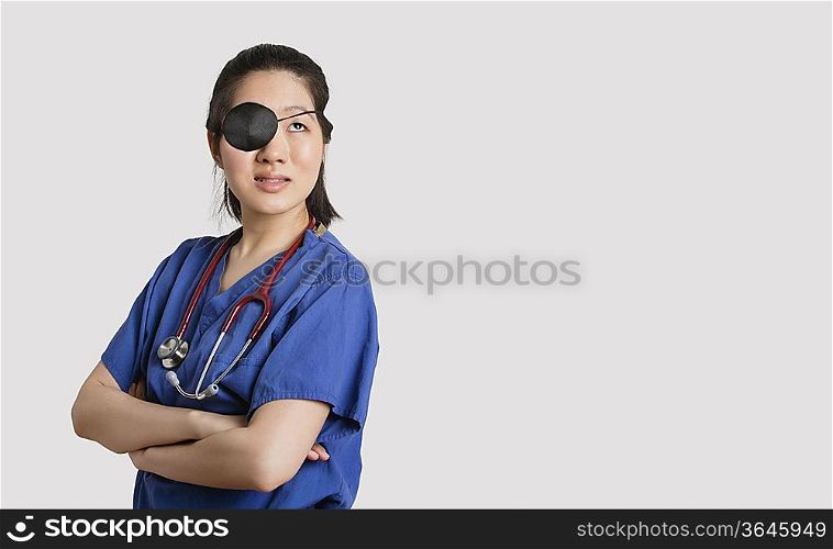 Asian female doctor wearing an eye patch looking up with arms crossed over gray background