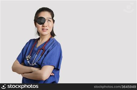 Asian female doctor wearing an eye patch looking up with arms crossed over gray background