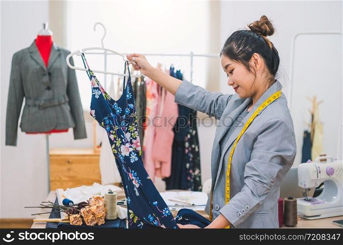 Asian female fashion designer girl making designing new beautiful blue dress clothes with hanger in studio. Fashion designer stylish showroom. Sewing and tailor concept. Creative dressmaker stylist.