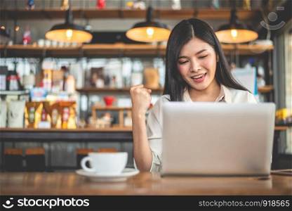 Asian female freelancer doing happy gesture by raising hand when using laptop in cafe. Business and success concept. Coffee shop and outdoors theme. Cafeteria background.