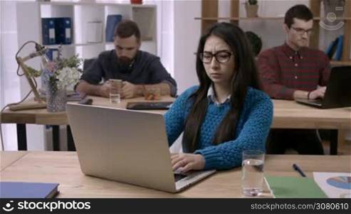 Asian female freelancer suffering from terrible headache at workplace and taking pills to relieve pain as her colleagues working on background. Sick businesswoman taking painkiller medicine at work.