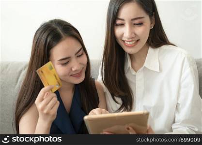 Asian female friends are using credit cards to order product online on tablets. While sitting on the sofa in the living room, Business transaction concepts