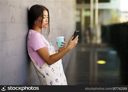 Asian female student taking a coffee break while consulting her smartphone. Chinese woman using a paper takeaway cup.. Asian female student taking a coffee break while consulting her smartphone.