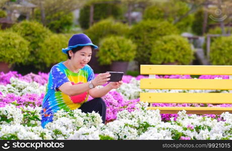 Asian female tourist in colorful casual style taking picture with smartphone in white and pink bougainvillea field on outdoor patio at Nong Nooch tropical garden, Thailand