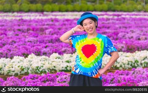 Asian female tourist in rainbow t-shirt is smiling cheerfully and looking at camera while posing in blooming pink and white bougainvillea field at Nong Nooch tropical garden, Thailand