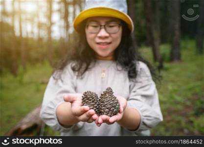 Asian female tourist in white dress with hat is holding pine cones in pine forest green on nature trail Forest Park.