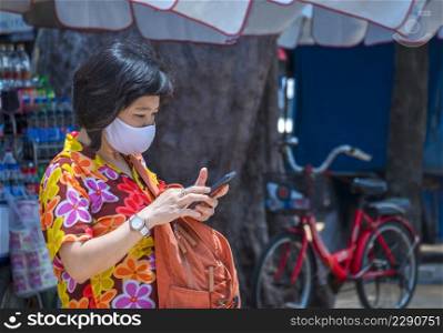 Asian female traveler in colorful casual style with protective mask using smartphone on walkway in seaside area