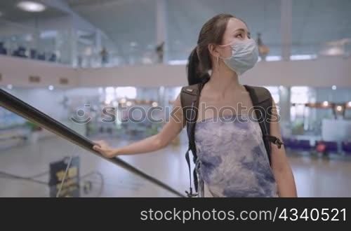 Asian female traveler wear protective face mask standing on escalator with backpack inside empty airport terminal, infectious diseases at public places hygiene, new normal pandemic, travel insurance