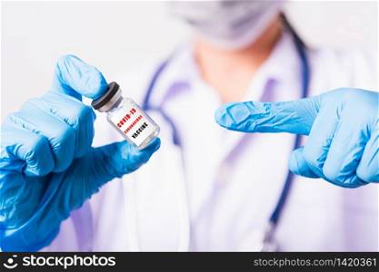 "Asian female woman doctor or nurse in uniform, gloves wearing face mask protective in laboratory pointing vial corona vaccine bottle on hand and bottle have "COVID-19 CORONAVIRUS VACCINE" text label"