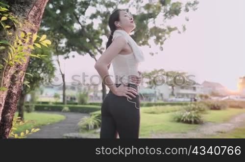 Asian fit young female stretching her waist lower back, warm up before exercise, body flexibility, health care, standing under the trees inside recreational exercise park, fresh morning work out