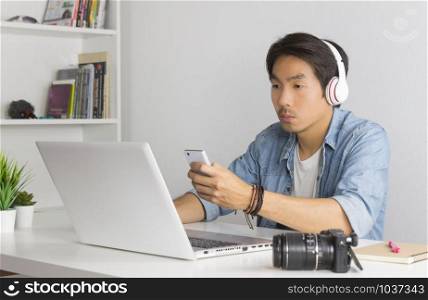 Asian Freelance Videographer in Denim or Jeans Shirt Checking Multimedia File by Smartphone with Laptop in Home Office. Freelance Videographer working with technology
