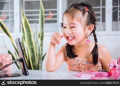 Asian funny little girl making makeup her face she look in smartphone and use eyebrow mascara, Learning activity to be woman, social media blogger recording vlog with cosmetics, online tutorial