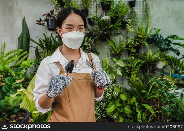 Asian gardener woman wearing face mask and apron stand and showing a shovel and pitchfork, equipment to transplants indoor plants and take care of plants at home, Hobby or small business with houseplant