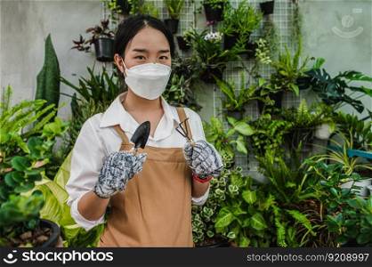 Asian gardener woman wearing face mask and apron stand and showing a shovel and pitchfork, equipment to transplants indoor plants and take care of plants at home, Hobby or small business with houseplant
