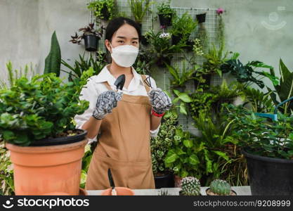 Asian gardener woman wearing face mask and apron stand and showing a shovel and pitchfork, equipment to transplants indoor plants and take care of plants at home, Hobby or small business with houseplant