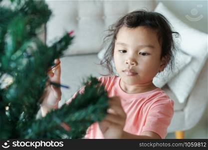 Asian girl are preparing a green Christmas tree for the holiday season in her home. The little girl is decorating the Christmas tree.
