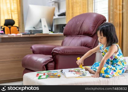 Asian girl child reading interactive book in living room at home as home schooling while city lockdown because of covid-19 pandemic across the world. Home Scholling concept.