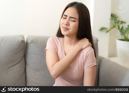 Asian girl having shoulder pain sitting on sofa at home. young girl has pain from the muscles, Health and illness concepts