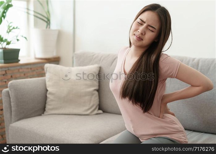 Asian girl having Waist pain sitting on sofa at home. young girl has pain from the muscles, Health and illness concepts