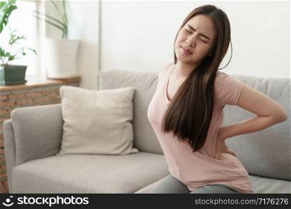 Asian girl having Waist pain sitting on sofa at home. young girl has pain from the muscles, Health and illness concepts